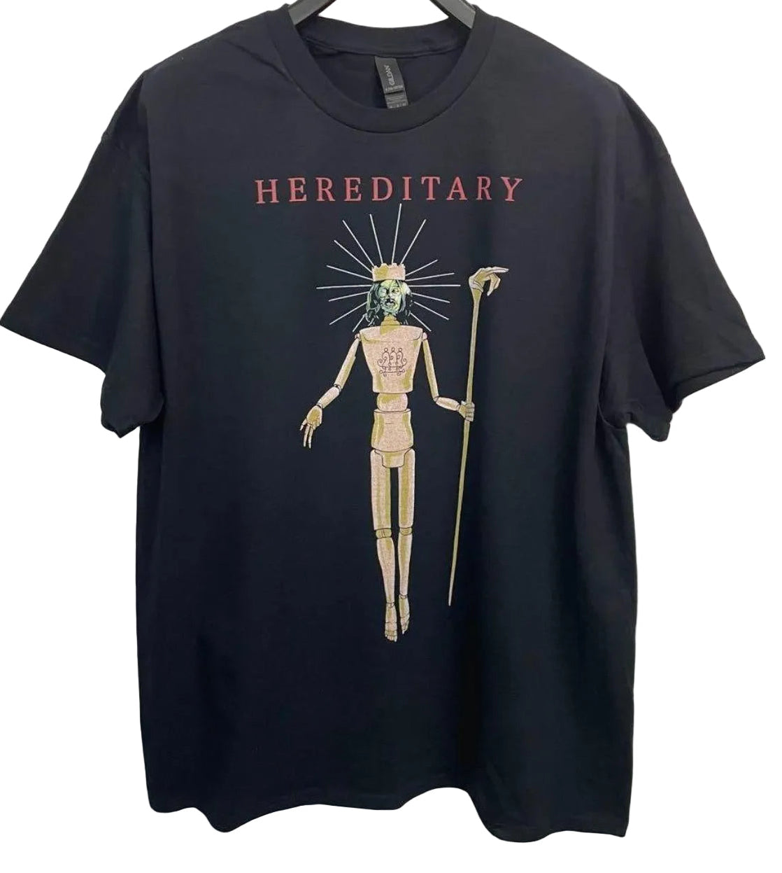 A black short-sleeve t-shirt hanging on a black hanger against a light gray wall. The shirt features a graphic print of a skeletal figure with a green head, holding a staff, with the word 'HEREDITARY' printed in red above the figure.